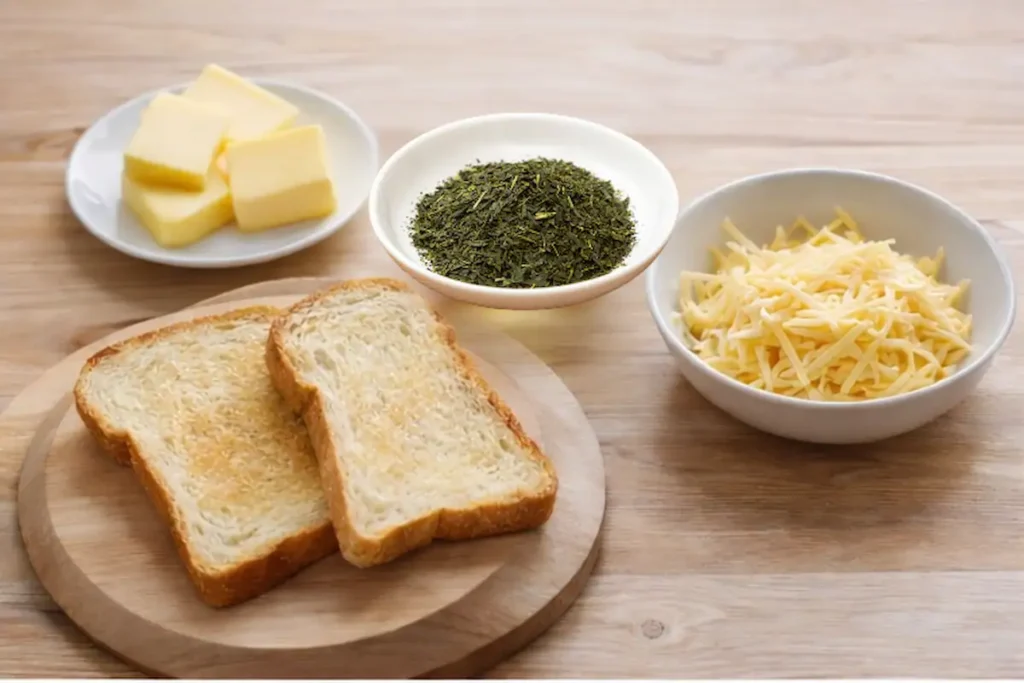Ingredients for cheese toast with green tea including bread slices, grated cheese, butter, and green tea on a wooden table